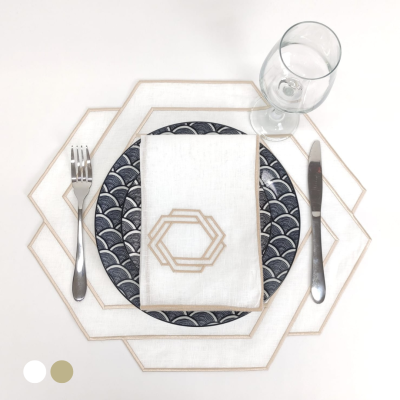 Jogo americano e guardanapo brancos com padrão dourado, prato azul e branco, talheres prateados e copo de vidro em mesa branca