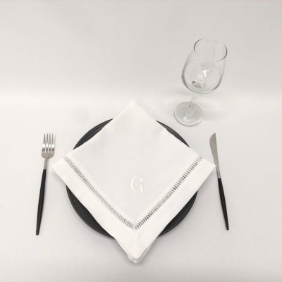 Mesa posta com prato preto, guardanapo branco bordado letra G, talheres com cabo preto e copo de vinho transparente.