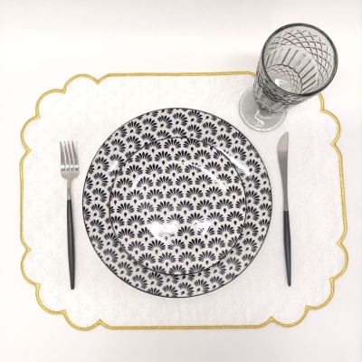 Conjunto de pratos brancos com padrão preto, talheres cinza e copo transparente sobre marcador de mesa branco com borda dourada