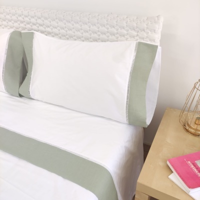 Fronha e lençol branco com detalhes verdes claros numa cama com cabeceira branca acolchoada e mesa de cabeceira com livros e lâmpada.