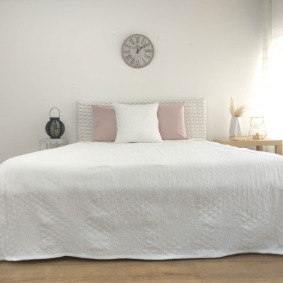 Quarto branco minimalista com cama de casal, edredão branco, almofadas rosa e branca, mesas laterais e relógio na parede