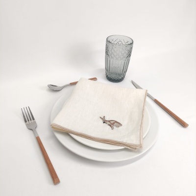 Mesa posta com talheres de madeira, prato branco, guardanapo com bordado de coelho e copo de vidro cinza.