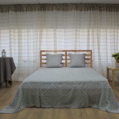 Quarto com cama de madeira, colcha cinza, cortinas brancas e mesas com decoração