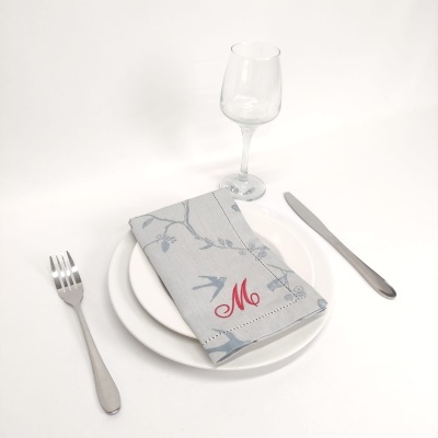 Conjunto de loiça com pratos brancos, guardanapo cinza com padrão e letra M vermelha, talheres e copo de vidro