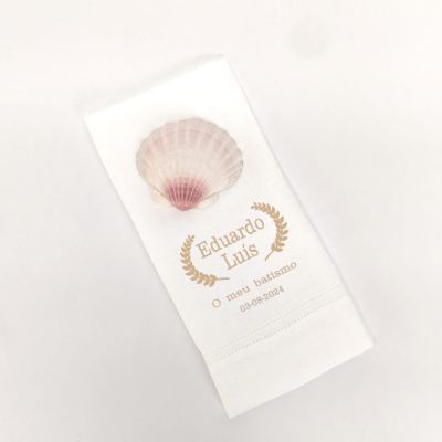Guardanapo branco com concha cor de rosa e texto dourado para batismo