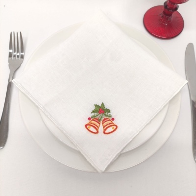 Guardanapo branco com bordado de sinos dourados e folhas verdes sobre prato branco com talheres e copo vermelho