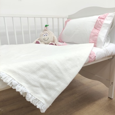 Cama de bebé branca com roupa de cama branca e cor-de-rosa, manta branca e peluche sorridente.