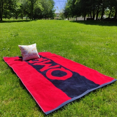toalha de praia vermelha e preta com texto OMK sobre relva com almofada branca