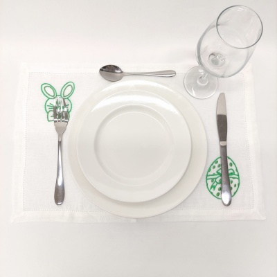 Jogo americano branco com bordados verdes, pratos brancos, talheres e taça de vidro