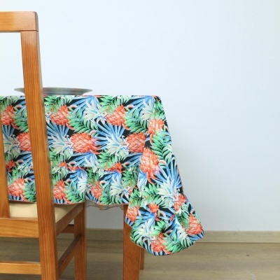 Toalha de mesa colorida com padrão de abacaxis e folhas de plantas sobre mesa e cadeira de madeira clara