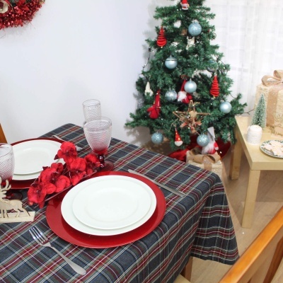 Mesa de jantar com toalha xadrez, pratos brancos e vermelhos, copos, flores vermelhas, árvore de Natal decorada e mesa com presentes