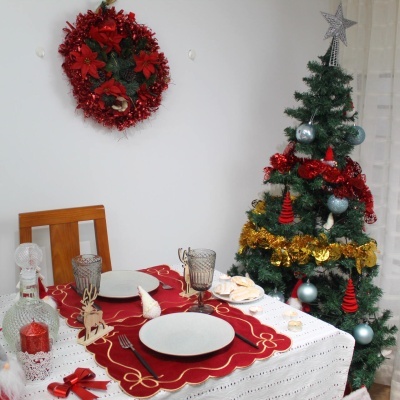 Mesa de jantar decorada para Natal com toalha branca e individuais vermelhos, ao lado de árvore de Natal decorada com bolas e fitas.