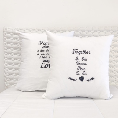 Duas almofadas decorativas brancas com bordados de texto preto sobre uma cama branca com cabeceira texturizada.