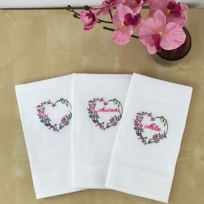 Toalhas brancas bordadas com corações florais e palavras para mãe e madrinha ao lado de flores cor-de-rosa em vaso.