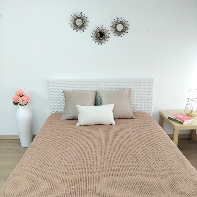 Cama arrumada com colcha bege e almofadas, vaso com flores e mesa de cabeceira com livros e vela