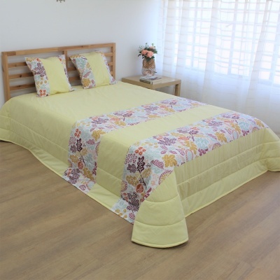 Cama de madeira clara com roupa de cama amarela e padrão floral, almofadas, mesa lateral com flores, cortinas brancas