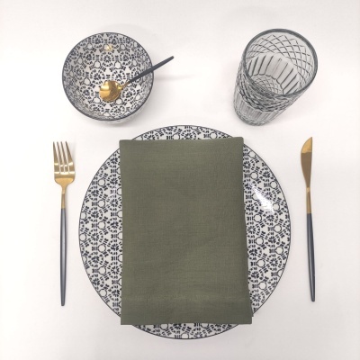 Conjunto de jantar com prato e tigela brancos com padrão azul, talheres dourados e pretos, guardanapo verde e copo de vidro com padrão geométrico