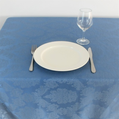 Mesa posta com prato branco, talheres em metal, copo de vidro e toalha azul com padrão floral