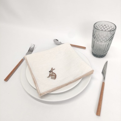 Conjunto de jantar com prato branco, guardanapo branco com bordado coelho, talheres de cabo madeira e copo de vidro padrão geométrico.