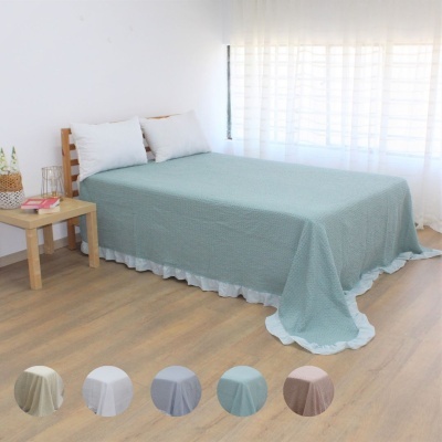 Cama solteiro com colcha verde água e almofadas brancas num quarto claro com mesa de madeira e cortina branca.