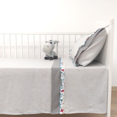 Cama de bebé com lençol cinza claro listrado, almofada cinza com borda ondulada padrão azul e vermelho, e peluche cinzento de burro