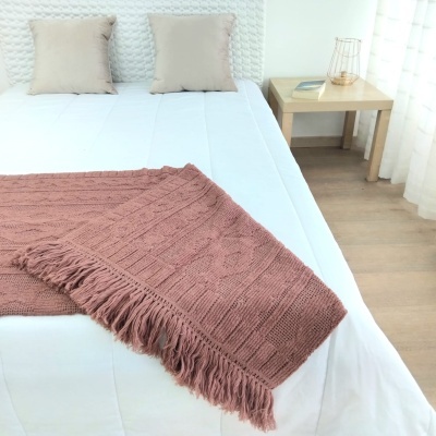 Cobertor ou manta rosa claro com padrão de tricô e franjas sobre cama branca com almofadas bege e mesa de apoio