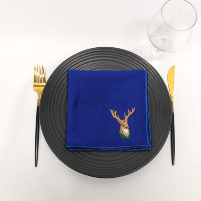 Guardanapo azul bordado com veado sobre prato preto com talheres dourados e copo transparente