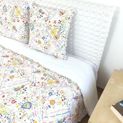 Cama com almofadas e edredão coloridos com padrões florais e texto, cabeceira branca texturizada e mesa de cabeceira com livros.