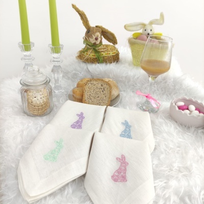 Guardanapos brancos com bordados de coelhinho em cores pastéis, com decoração de páscoa e pães sobre mesa felpuda branca