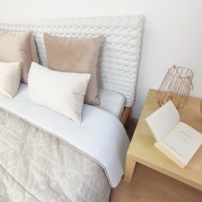 Cama com almofadas bege e branca e mesa de cabeceira com livro aberto e candeeiro dourado