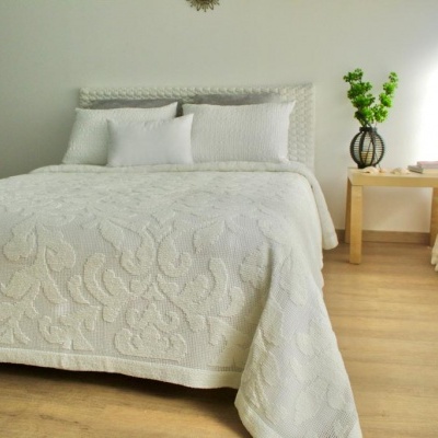 Cama com colcha branca texturizada com padrão floral, almofadas brancas, mesa de madeira com vaso e cortinas brancas