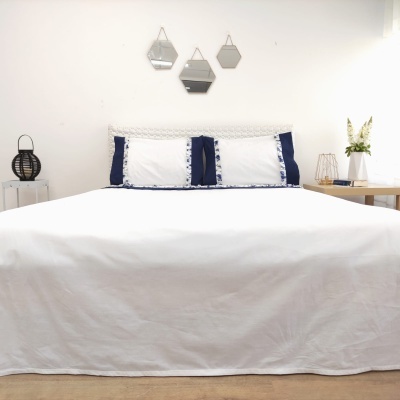 Cama com colcha branca e almofadas brancas com bordas azuis, espelhos geométricos na parede e decoração simples ao redor.