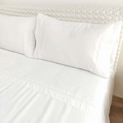 Lençóis e fronhas brancos com bordado decorativo numa cama com cabeceira branca texturada