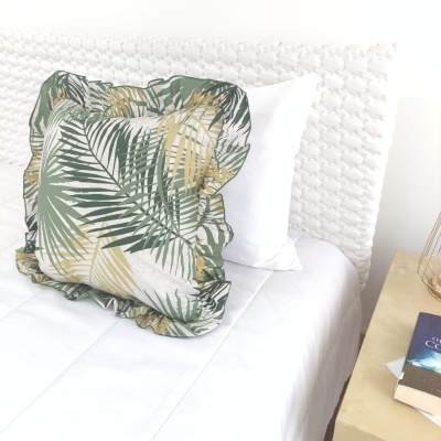Almofada decorativa com padrão de folhas sobre cama branca com cabeceira texturizada e mesa de cabeceira com livros
