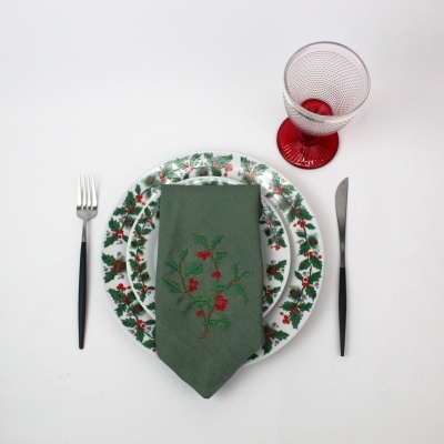 Conjunto de louça com padrão de folhas e frutos vermelhos, guardanapo verde bordado, talheres e copo vermelho texturizado.