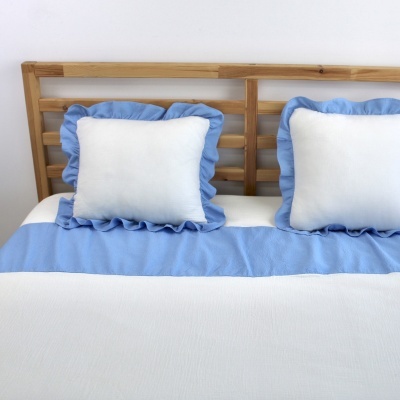 Cama com colcha branca e azul claro e almofadas com folho azul claro sobre cabeceira de madeira clara