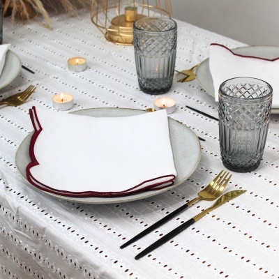 Mesa posta com pratos, guardanapos brancos com borda vermelha, copos cinza texturados, talheres preto e dourado, toalha branca com furos, velas e decoração.