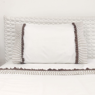 Almofada branca com laterais riscadas cinza e guarda castanha numa cama com cabeceira branca acolchoada