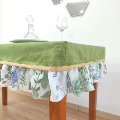 Capa de mesa verde com saia floral e acabamento em renda bege sobre mesa de madeira