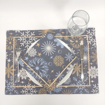 Jogo americano e guardanapo com padrão de flocos de neve azul, branco e dourado, prato branco, talheres de metal e copo transparente sobre mesa branca