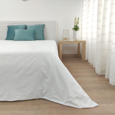 Cama branca com almofadas azuis, mesa lateral de madeira com flores e luminária e cortinas brancas