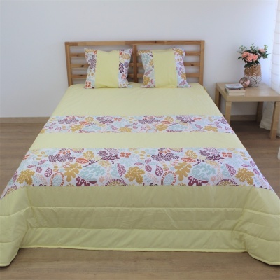 Cama com colcha amarela clara com motivos florais e duas almofadas; cabeceira de madeira clara; mesa de cabeceira com livros e vaso de flores.