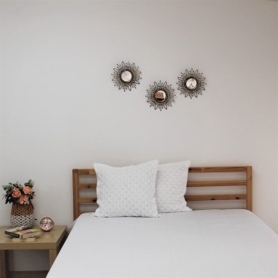 Quarto com cama de madeira, almofadas brancas, mesa de cabeceira com livros, relógio e flores, e três espelhos decorativos na parede branca.