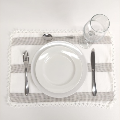 Mesa posta com prato branco, talheres prateados, copo transparente e individual branco com listra cinza e bordado