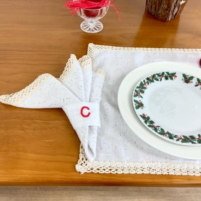 Jogo americano e guardanapo branco com renda bege e porta-guardanapo com letra C, prato com padrão floral.