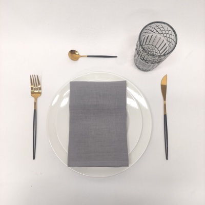 Conjunto mesa com prato branco, guardanapo cinzento, talheres preto e dourado, copo transparente com padrão geométrico
