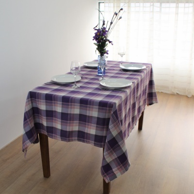 Mesa posta com toalha xadrez roxa e lilás, pratos, copos e vaso com flores