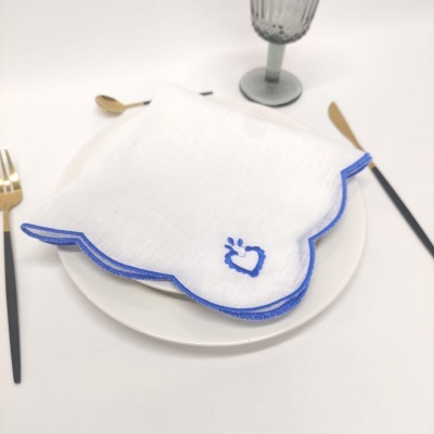Guardanapo branco com borda azul e bordado azul sobre prato branco, talheres dourados e cabo preto, copo cinza texturizado