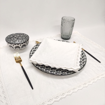 Mesa posta com louça floral a preto e branco, talheres preto e dourado, copo em vidro verde-fumado, e toalha branca bordada