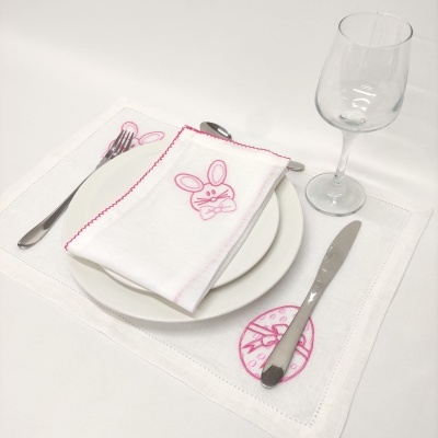 posto de mesa branco com bordado rosa de coelho, talheres metálicos e copo de vidro